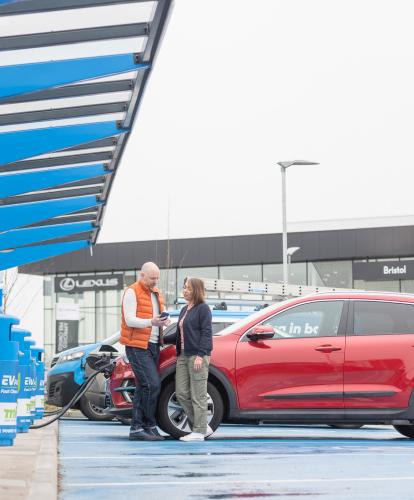 Couple stand in front of charging EV, Car dealership in background