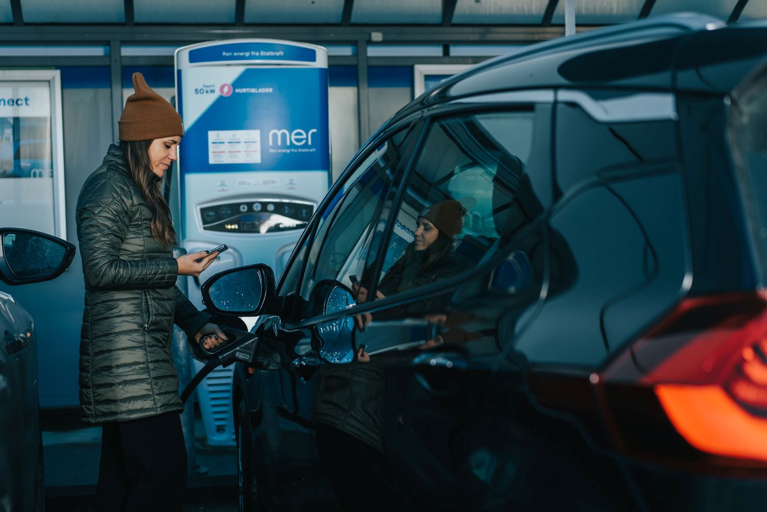 Mer charger - lady with a brown hat chargers her EV while holding phone