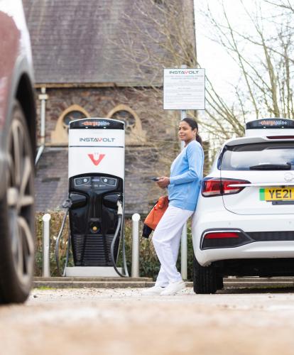 Woman leans on EV looking at Zapmap app on phone while EV charges