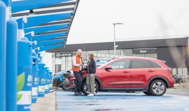 Couple stand in front of charging EV, Car dealership in background