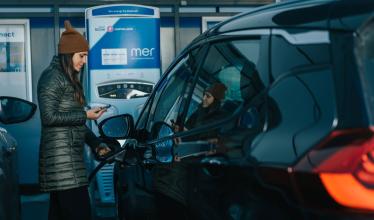 Mer charger - lady with a brown hat chargers her EV while holding phone